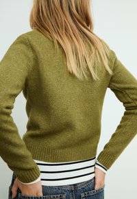 Olivenfarvet strikket sweater med ribbet kant og manchetter, med en rund halsudskæring. Stribet hvid og sort skjorte lagdelt nedenunder.