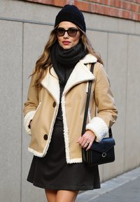 Giacca in ecopelle beige con bordo in shearling bianco, dolcevita nera, gonna scura, sciarpa nera e borsa; indossata con un berretto di maglia nero.