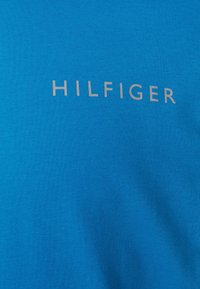 Baumwoll-T-Shirt in leuchtendem Blau mit einer glatten Textur, auf der die Markenbezeichnung "HILFIGER" in silbernen Buchstaben auf der Vorderseite prangt.