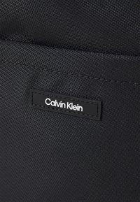 Crna platnena torba s glatkom teksturom, štepanim detaljima i pravokutnom crnom etiketom s natpisom "Calvin Klein" u bijelom slovima.