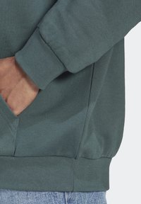 Handen i fickan på en mörkgrön sweatshirt med ribbad mudd och midja, i kombination med ljusblå denimjeans.