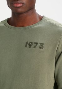 Olivgrön bomulls skjorta med lång ärm, ribbad halsringning och broderad "1973" på bröstet, med en mjuk textur och avslappnad passform.