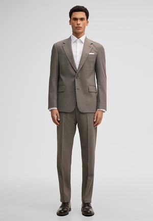 Mann steht und trägt einen braunen, maßgeschneiderten Anzug mit Kerbkragen, weißem Hemd, weißem Einstecktuch und dunkelbraunen Anzugschuhen vor einem schlichten Hintergrund.