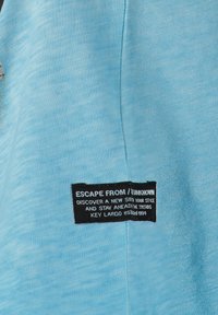 Tejido de color azul claro con una superficie texturada. Presenta una etiqueta negra con texto blanco que muestra detalles de la marca y el eslogan.