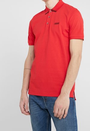 Hombre con camiseta polo roja de manga corta con ribete negro y vaqueros azules, mostrando los antebrazos tatuados y las manos relajadas a los costados.