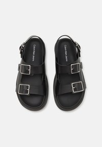 Svarta sandaler i gummi med två breda remmar som har silverfärgad spännen. Innersulorna är släta med varumärket tryckt i vitt.