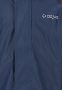 Marinblå vattentät jacka med en slät yta, utrustad med en vit zigzag-logotyp och kontrasterande mörka detaljer på axlarna.
