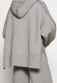 Przytulny, oversize szary hoodie z miękkiego materiału, z kapturem, opuszczonymi ramionami i czarnymi przeszyciami wzdłuż szwów, prezentowany od tyłu.