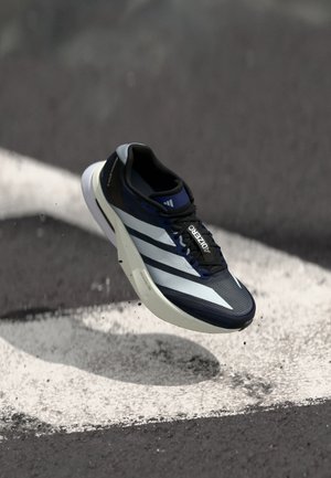 Niebiesko-biały but do biegania Adidas Adizero unoszący się w powietrzu nad asfaltem z białą, zakrzywioną linią oznaczenia, widoczne małe zanieczyszczenia wokół podeszwy.