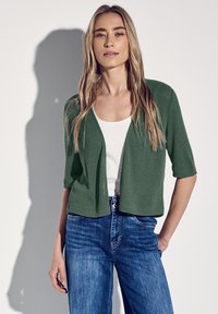 Cardigan vert ouvert à l'avant avec des manches courtes, associé à un haut blanc et un jean bleu. Tissu en maille texturé. Design simple, coupe décontractée.