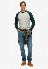 Mand iført en grønærmet raglantrøje, blå jeans, brune læderstøvler, en ternet skjorte bundet omkring taljen og et armbåndsur, der står med hænderne i lommerne.