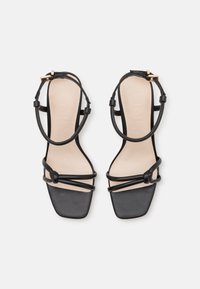 Sandalias de tacón alto negras con punta cuadrada. Presenta tiras delgadas y un detalle anudado. La suela es de color nude y ligeramente acolchada. Tiene un acento de hardware dorado.