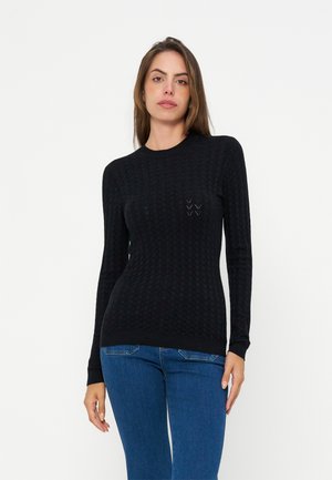 Femme aux longs cheveux bruns portant un pull noir texturé à manches longues et un jean bleu, debout devant un fond blanc uni.