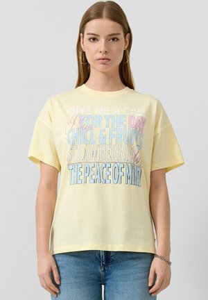 Jeune femme aux cheveux raides portant un t-shirt jaune pâle avec un texte multicolore et un jean bleu, debout devant un fond gris clair.