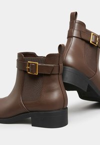 Botas de tobillo de cuero marrón con un panel texturizado, acento de hebilla dorada y un bajo tacón de goma negra. Elástico lateral para facilitar el uso.