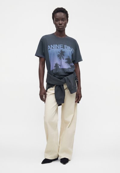 Γυναίκα που φοράει μαύρο γραφικό T-shirt με φοίνικες, μπεζ παντελόνι με φαρδιά μπατζάκια, μαύρα μυτερά παπούτσια και μαύρο φούτερ δεμένο στη μέση.