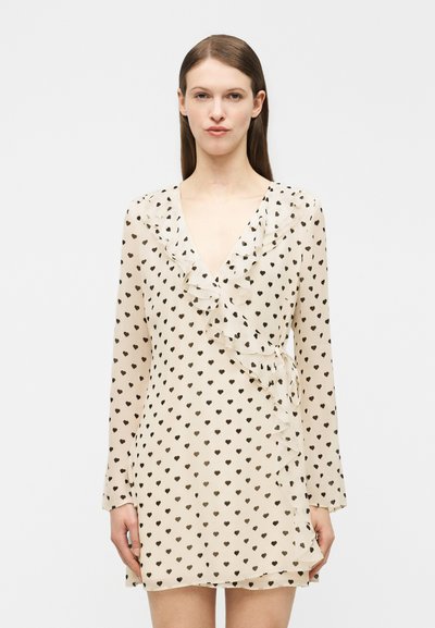Femme portant une robe portefeuille blanche transparente avec des motifs de cœurs noirs et des volants, debout devant un fond clair uni.
