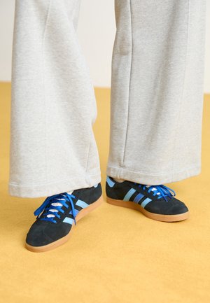 Czarno-niebieskie sneakersy z gumowymi podeszwami noszone z jasnoszarymi szerokimi spodniami na żółtej podłodze.