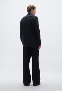 Sötét hosszú ujjú ing klasszikus gallérral, széles szárú fekete nadrággal párosítva. Az anyag texturált hatású, rétegzett megjelenést kölcsönözve.