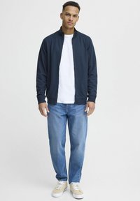 Tengerészeti zipzáras dzseki magas gallérral, alatta fehér póló, világoskék farmernadrág és bézs sportcipő fehér fűzővel.