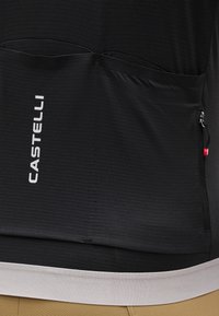 Κοντινό πλάνο της πίσω τσέπης μαύρης ποδηλατικής φανέλας Castelli με μικρό φερμουάρ και λευκή κάτω ραφή, πάνω από καφέ σορτς.