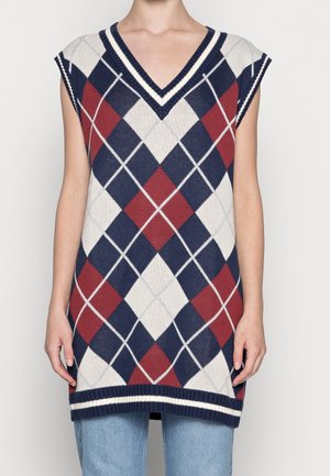 Bezrekawnik z dzianiny z granatowo-czerwono-białym wzorem w romby i dekoltem w serek, noszony na jasnoniebieskich dżinsach.
