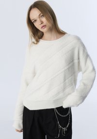 Pull blanc texturé avec des motifs en côtes diagonales, décolleté large et ourlet côtelé. Associé à un pantalon noir agrémenté de chaînes.