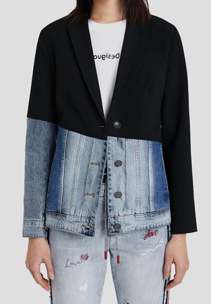 Femme portant un blazer noir associé à une veste en denim bleu clair et un jean bleu clair avec des textes et des motifs brodés.