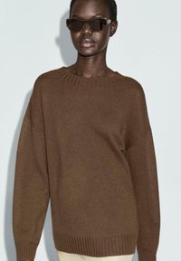Persona di pelle scura che indossa un ampio maglione di maglia marrone e grandi occhiali da sole angolari scuri, in piedi contro uno sfondo grigio chiaro.