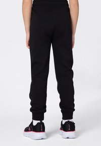 Persoon die staat en zwarte joggerbroeken en zwarte sneakers met witte zolen en roze accenten draagt, gezien van achteren.