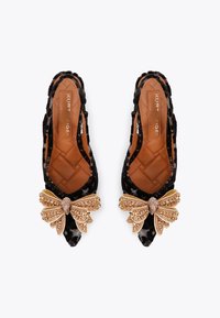 Flats en velours noir avec un grand accent de nœud en bijoux de tons dorés, semelle intérieure matelassée et design à dos ouvert. Texture lisse avec des détails élégants.