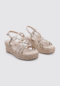 Sandali con zeppa dotati di una suola in fibra naturale, dettagli circolari metallici e una tomaia beige chiaro. Design con punta aperta e cinturini alla caviglia.