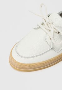 Chaussure blanche en cuir avec une finition texturée, bout arrondi et semelle en caoutchouc beige. Comprend des lacets blancs et des détails de couture bleu clair.