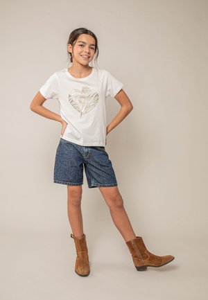 Glimlachend meisje staat met haar handen in haar heupen, draagt een wit t-shirt met een gouden hart, spijkershort en bruine suède enkellaarsjes.