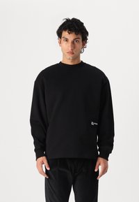 Sweatshirt negru cu guler rotund și mâneci lungi, având un detaliu cu logo în partea stângă jos. Fabricat dintr-un material moale și texturat.