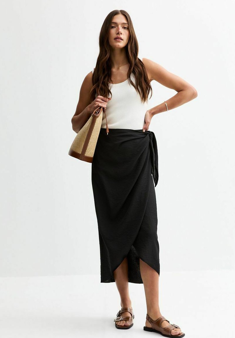 Jupe enveloppante noire avec une fente haute, portée avec un débardeur côtelé blanc. Le modèle porte un sac en paille tissé avec des accents bruns, assorti à des sandales brunes.