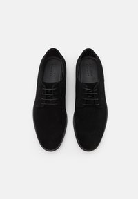 Scarpe in suede nero con texture liscia, punta arrotondata e dettagli di laccio sottili; design minimalista e suola piatta.