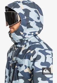 Blå og grå jakke med camouflagemønster, højt krave, justerbar hætte og sort logo-patch. Glat tekstur, letvægtsmateriale.