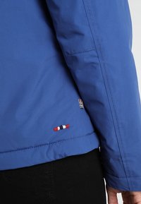 Modrý rukáv bundy a spodní část zad s malým vyšívaným detailem v červené, bílé a modré barvě, nošeno přes černé kalhoty.