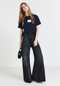 Donna con lunghi capelli biondi, che indossa una maglietta blu navy e pantaloni neri a gamba larga, con in mano una piccola borsa nera, in piedi davanti a uno sfondo bianco.