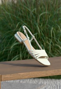 Sandale blanche à bride arrière avec petit talon carré, présentée sur une surface en bois devant un fond d'herbes hautes vertes.