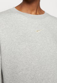 Szary sweter z miękką fakturą, z małym haftowanym złotym logiem Nike na piersi. Prosty dekolt typu crew neck.