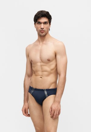 Muskelmand står iført marineblå badebukser med hvide striber, foran en ensfarvet lysegrå baggrund.