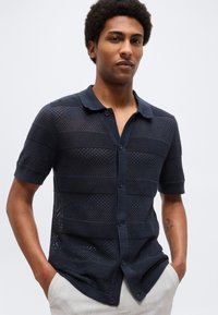 Άνδρας με μπλε σκούρο κοντομάνικο πουκάμισο από δίχτυ με γιακά, στέκεται με τα χέρια στις τσέπες ανοιχτόχρωμων παντελονιών.