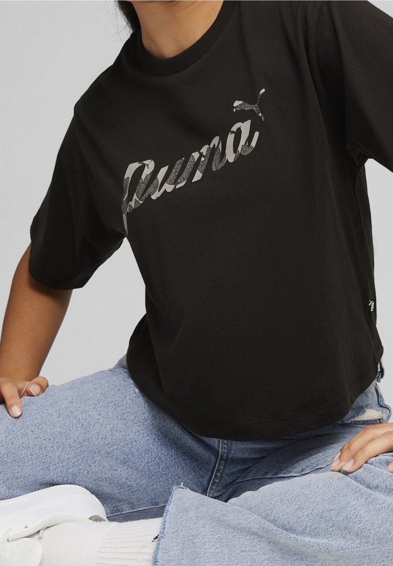 Personne portant un t-shirt noir Puma avec un logo gris, un jean bleu clair, des chaussettes blanches et des baskets blanches, assise les jambes croisées.