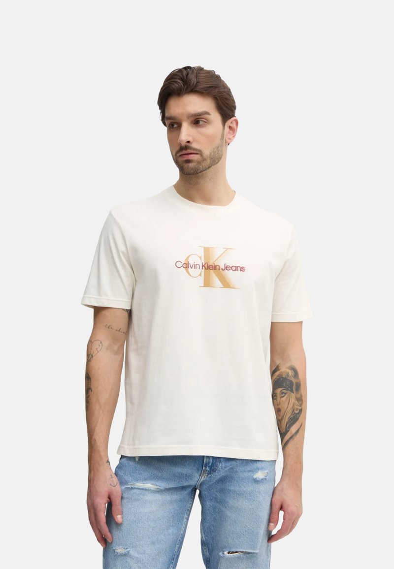 Camiseta de algodón blanca con un audaz logo de Calvin Klein en naranja y burdeos. Mangas cortas, corte relajado y tatuajes visibles en el brazo del modelo.