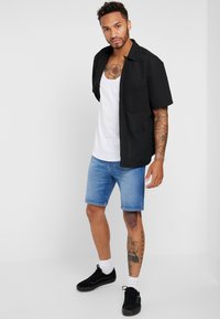 Sort kortærmet skjorte med en lomme, hvid top, blå denimshorts og sorte sneakers med hvide sokker; tatoveringer synlige på armen.