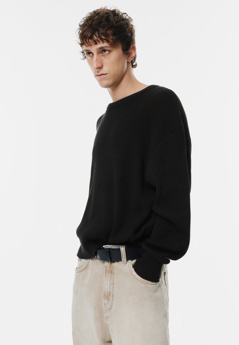 Maglione a coste nere con scollatura ampia, abbinato a pantaloni beige chiaro. Il modello sta in posizione casual, con le mani in tasca.