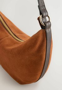 Sac bandoulière en daim marron avec une sangle en cuir foncé et un détail d'anneau métallique doré, avec une fermeture éclair visible sur le dessus.