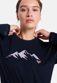 Marineblauw sportshirt met een lichtpaarse bergafbeelding op de voorkant en een klein roze logo. De stof lijkt zacht met een gladde textuur.